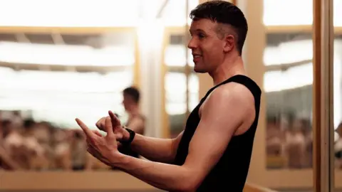 Daniel Greenan A man is teaching a yoga class. He has dark hair and is wearing a black vest. The room he is sitting in has a number of mirrors. 