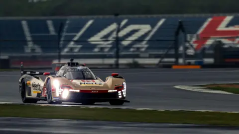 Jack Valpy/BBC The gold Cadillac Hertz Team JOTA Hypercar races through a sweeping bend at Silverstone Circuit, with bright headlights cutting through overcast conditions and empty grandstand seating blurred in the background.