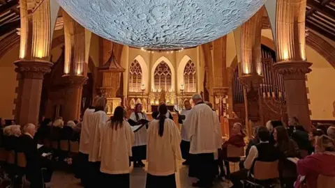 Cathedral Isle of Man A close-up of the bottom half of the suspended moon, which has visible craters, with the arches of the cathedral on either side and choristers underneath.