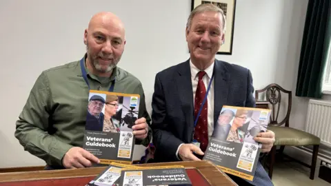 Two men sit at a table in a council office, facing the camera and holding copies of a handbook titled Veterans' Guidebook, which includes images of armed forces personnel on the cover