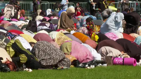 Muslims in colourful clothes kneeling and performing their prayers on a grassy field, all facing in the same direction