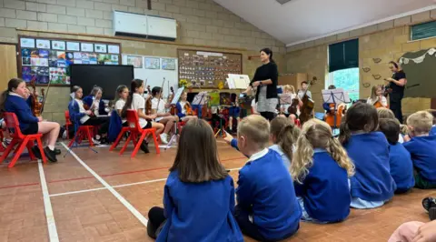School pupils holding instruments sit on plastic chairs in a school hall, with music stands in front of them. A row of pupils wearing blue school jumpers sit on the floor watching them.