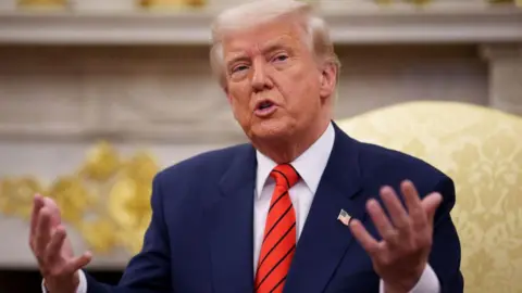 Donald Trump in the Oval Office, sitting and talking, with both hands held out in front of him. He is wearing a navy blazer with a US flag pin on his left lapel, a white shirt and a red and navy striped tie.