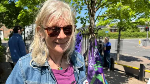 A smiling Lynne Hubbard at the protest in Dorchester. It's a sunny day. She is wearing sunglasses and has shoulder length blonde hair. She is wearing a denim jacket and a cerise t-shirt.