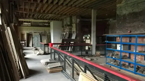 The dingy inside of an upstairs room at Ashton Court mansion showing doors stacked against a far panelled wall, bare floors and stripped ceilings with exposed beams. There are industrial red and black banisters in the centre of the picture, and blue industrial shelving holding building materials to the right.