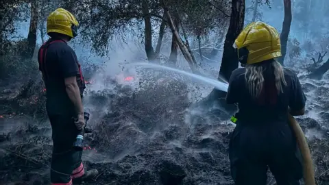Peterborough Volunteer Fire Brigade Smoke rises from the blackened floor, as a woman firefighter with long blonde hair shoots water at glowing embers, while a man watches on.
