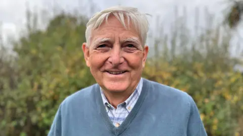 A man with white hair and a blue jumper standing in front of green bushes