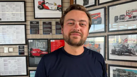 A close-up of a man wearing a navy T-shirt with the word “Takona” printed in white. He is standing indoors in front of a wall covered with framed motorsport posters, race diagrams and images of Formula 1 cars.