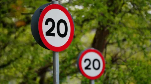 Two round 20mph speed limit signs against a backdrop of green trees - one sign in the foreground and one further away and out of focus