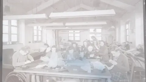 South West Heritage Trust A negative shows residents of St Loye's College and School of Occupational Therapy, taking part in a sewing workshop. It is hard to distinguish everything in the image but women or girls are sewing by hand and one can be seen sitting in front of a sewing machine. To the left of the photo a wheelchair user is working on a dark piece of fabric.