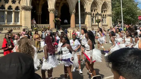 Carroll Weston/BBC People wearing white tops with fluffy white skirts dancing in front of a grand civic building.