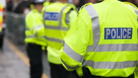 Three police officers standing outside with their backs to the camera. They are all wearing fluorescent yellow jackets with silver stripes along the arms and backs. On the back of each of their jackets in a blue rectangle and in grey writing it says POLICE.