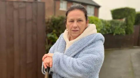 Janet Brooks holding a mop wearing a blue dressing gown stood outside her flooded property looking disgruntled. 