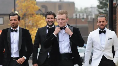 Getty Images TOWIE stars wearing tuxedos walking along a residential street to Sugar Hut. All tuxedos, completed with bow ties, are black with the exception of Pete Wicks, whose jacket is white. They all appear to be having a laugh together.