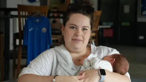 SHAUN WHITMORE/BBC Charlotte Robinson holding baby Henry in her arms. He is just two weeks old and is wearing a cream coloured bodysuit. Henry has brown hair and charlotte is wearing a smartwatch on one hand with a silver bracelet on the other. She is wearing a white top and has her dark brown curly hair up in a bun.