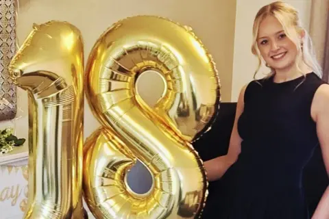 Helen Draper Meg, a teenage girl with long, curly, blonde hair wearing a black dress, smiling and looking at the camera. She is stood next to large gold balloons in the shape of the number 18.