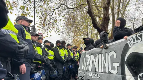 Police line in front of counter-protesters at Saturday's demonstration. The protesters are all wearing black with covered faces and sunglasses. Officers are wearing high vis yellow jackets on black uniforms.