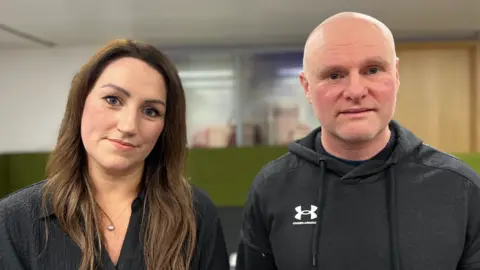 A woman with long hair is on the left next to a man. They are both wearing dark clothing.