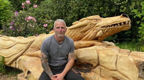 Scott Elson sits on a fallen lime tree which has been carved into the shape of a dragon. It sits among woodland.