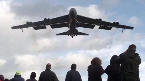 A B52 Stratofortress bomber flying out of RAF Fairford. There is a group of people below watching the plane take off.