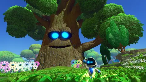 Sony A small robot with a white body, black screen for a face and two bright blue eyes frolics in a green field full of colorful, identical flowers arranged in neat rows. Behind him, a giant tree with a smiling face and bright blue eyes looks over the jolly scene.
