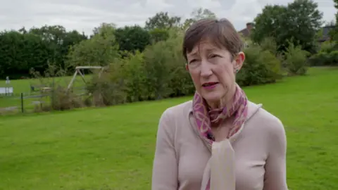 A woman with cropped brown hair, a light pink jumper, a pink patterned scarf, blue stud earrings and pink lipstick stands in a park.