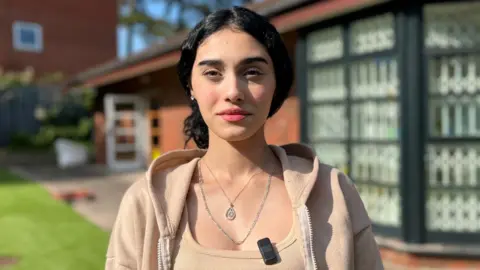 A girl with dark hair is wearing a beige jacket and top. She is standing in front of a brick building.