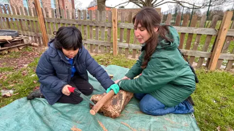 Martin Giles/BBC A pupil and teacher outside cutting a wooden branch. The pupil, on the left is holding a small saw, has dark hair and is wearing a navy coat. The teacher to the right is holding the branch with both hands and is wearing a green coat and blue jeans.