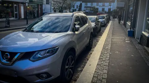 An SUV parked on a residential street. the silver car is in front of a row of shops with white tudor style Maltsters pub in the background. 