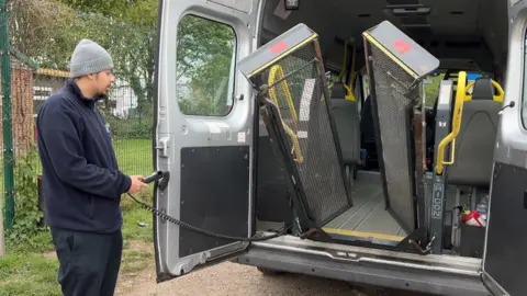 A private hire operator, wearing a wooly hat, uses a handset to lower a lift used to get wheelchairs into the back of a specially adapted minibus.