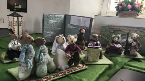 Eleven knitted mice at a staged mouse wedding with a photo of Jurby in the background and mini sculpture church.