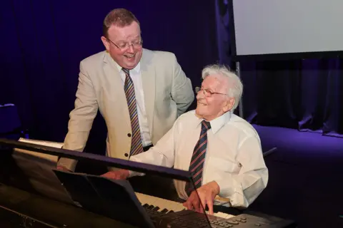 The Croft Mr Bickley sits at the organ with his hands on the keys and smiles. Mr Lloyd stands next to him and grins at him. Mr Bickley is wearing a white shirt and blue and red striped tie, while Mr Lloyd wears a cream suit jacket, white shirt and striped tie