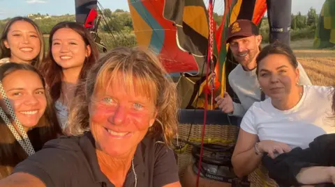 Six people standing in a balloon basket on a hot sunny day with a hay field in the background. The balloon pilot in the centre of the picture has mid length mousy blonde hair and wearing a short sleeved dark coloured shirt.