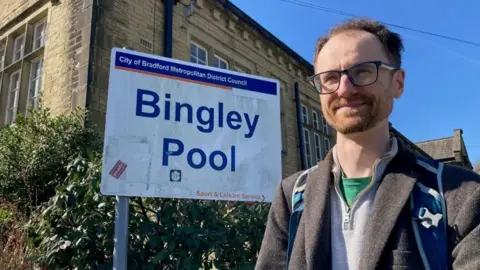 Charles Heslett/BBC A man with short curly hair and beard wearing rectangular glasses stands next to a sign reading 'Bingley Pool'