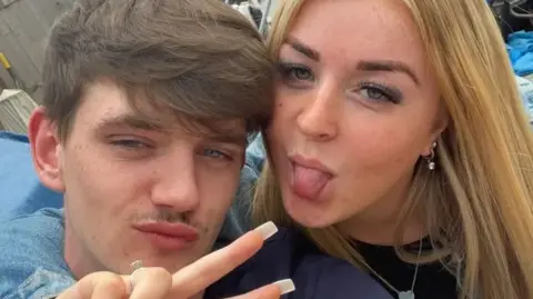 Tyler Kerry, who has brown hair swept across his head and is pouting. A woman with blonde hair has her arm around him and is doing a peace sign with her fingers while sticking her tongue out.
