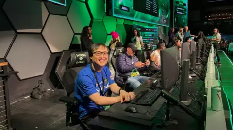 Ha Dang Ha Dang smiles at the camera. He is sitting in front of a computer alongside a row of several computers and other people on a stage. Behind him are green lights and a large screen showing the results of the competition.