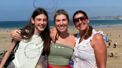 Family handout Ms Exton with her daughters Jess and Sophia stood arm in arm in front of a beach and the ocean.