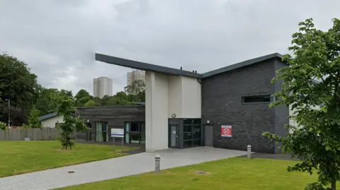 A path leads to a modern hospital unit with a sloped roof hanging over a single-storey section clad in wood on the left of the glass doors and a grey brick two-storey section to the right. There is a sign saying 'Syke House'.  The unit is flanked by trees and two high-rise tower blocks can been seen in the distance. 