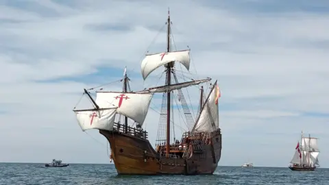 The Nao Victoria Foundation A wooden ship in the ocean. It has large white masts with a red gross on them. There is a Spanish flag flying from the back.