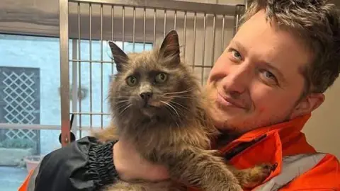 Sue Vidamour A long-haired cat with brown fur is very alert as she is held by her smiling owner  who is dressed in an orange jacket.