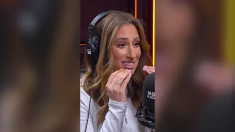 Woman with brown hair and headphones on her head holds her hands near her mouth.