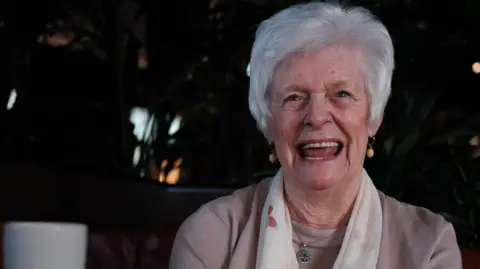 BBC Marie McLaren smiles for the camera. She has short grey hair and is wearing a white scarf.
