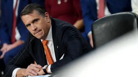 Todd Blanche wears a balck suit with an orange tie. He is seen sitting at a desk at a Senate confirmation hearing, with the frame tilted towards the left so the image appears at an angle.
