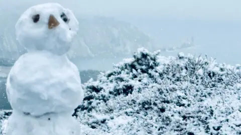 A snowy scene from a clifftop above a beach. A small snowman has been made with three items places on its head to make eyes and a nose.