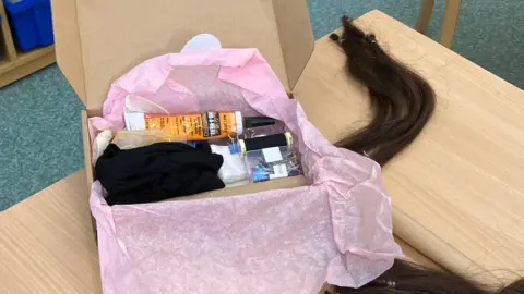 A box containing all the equipment needed to make a hat of joy. It contains an orange tube of glue, a box of pins, black thread and pieces of black and white material. There is pink tissue paper in the box and it is placed on the table. 