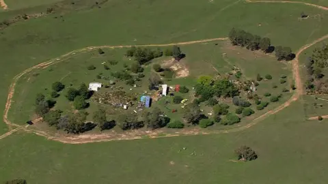 Australian Broadcasting Corporation An aerial view of a grassy area with containers in the middle surrounded by trees and a mud track