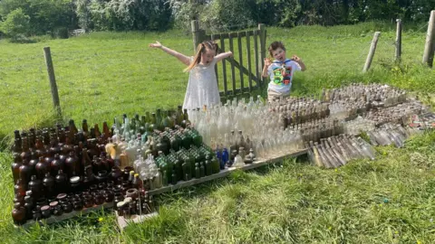 Zoe Brown The entire pile of bottles with two children standing behind them. A little girl with blonde hair and a white dress is holding her arms above her head and the little boy in a white t-shirt is holding something in his hands. There are rows and rows of clear, green and brown bottles of different shapes and sizes in front of them.