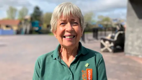 Sally Everton is pictured smiling. She is wearing a green polo shirt with the Visit Devon logo on it.