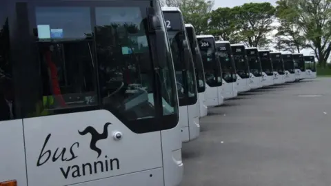 IOM GOVERNMENT A row of the fronts of modern silver single-decker buses. The Bus Vannin logo, which features the the three legs of Mann can be seen on the side of the bus at the head of the line.