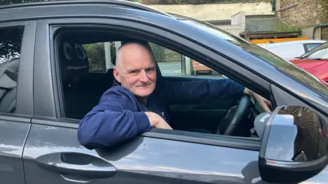 An older man leans out of the window of a car, smiling